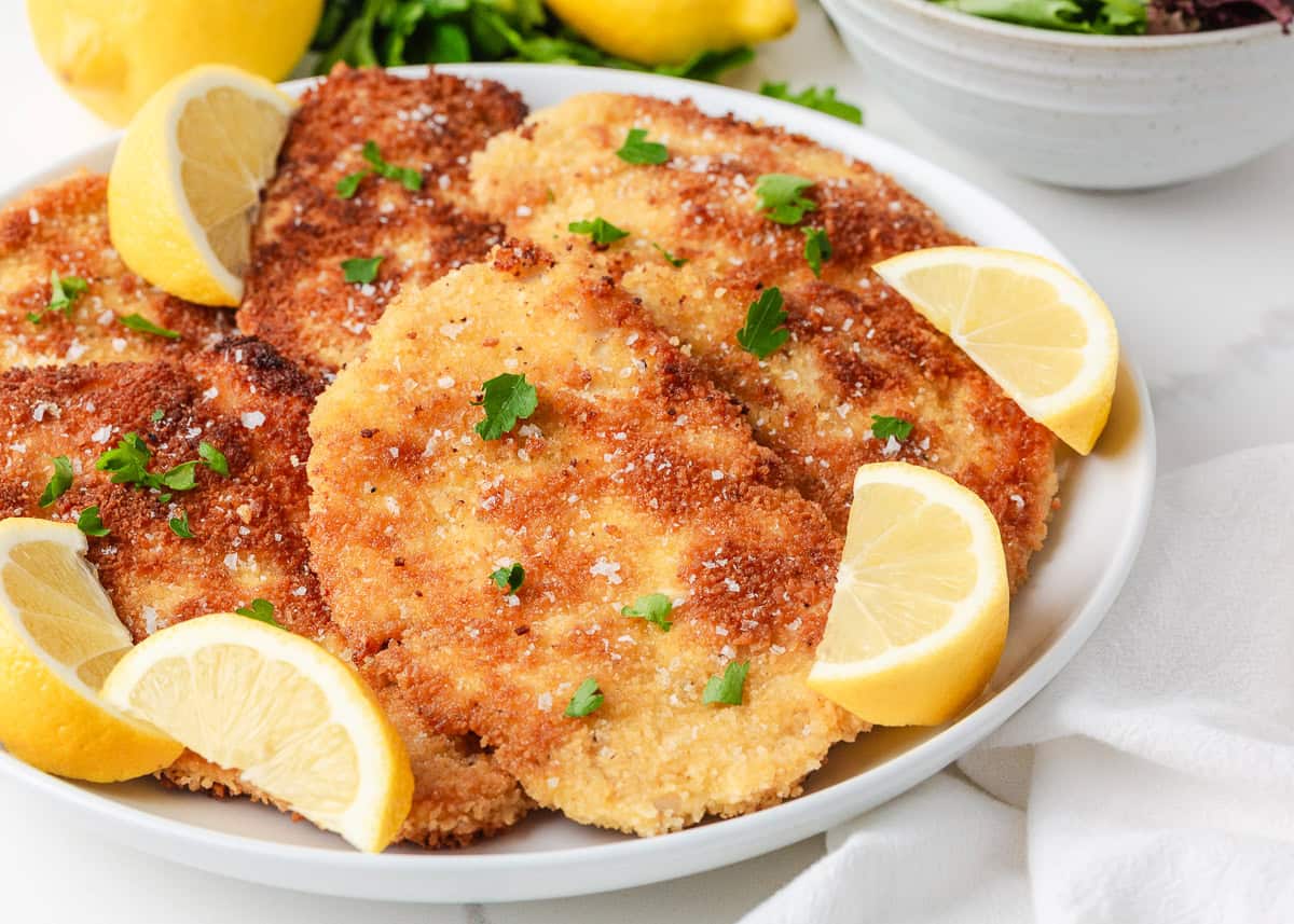 This chicken schnitzel is coated in seasoned breadcrumbs and pan fried until perfectly crispy, then finished with a squeeze of lemon. Thin, juicy, and ready in 30 minutes! Crispy pan fried chicken schnitzel cutlets arranged on a plate with fresh parsley and lemon.
