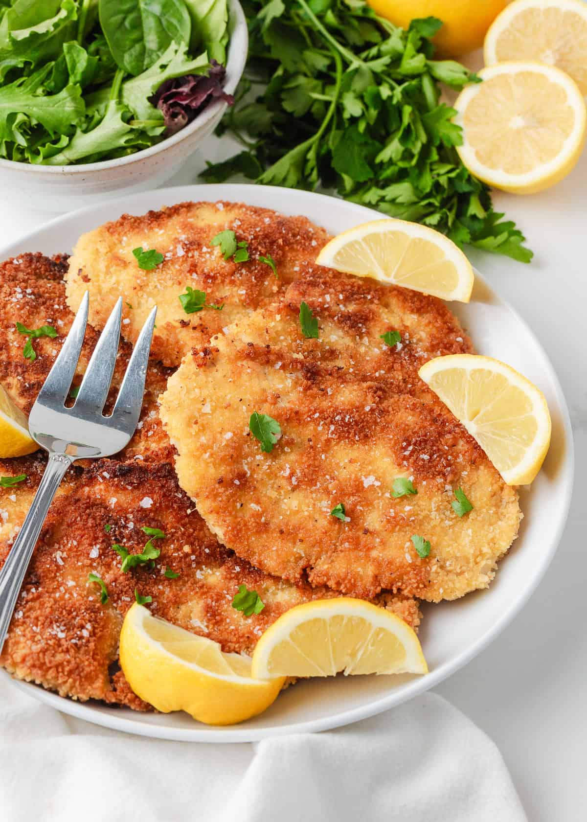 This chicken schnitzel is coated in seasoned breadcrumbs and pan fried until perfectly crispy, then finished with a squeeze of lemon. Thin, juicy, and ready in 30 minutes! Crispy chicken schnitzel cutlets on a white plate, topped with flaky salt and parsley and served with fresh lemon wedges.