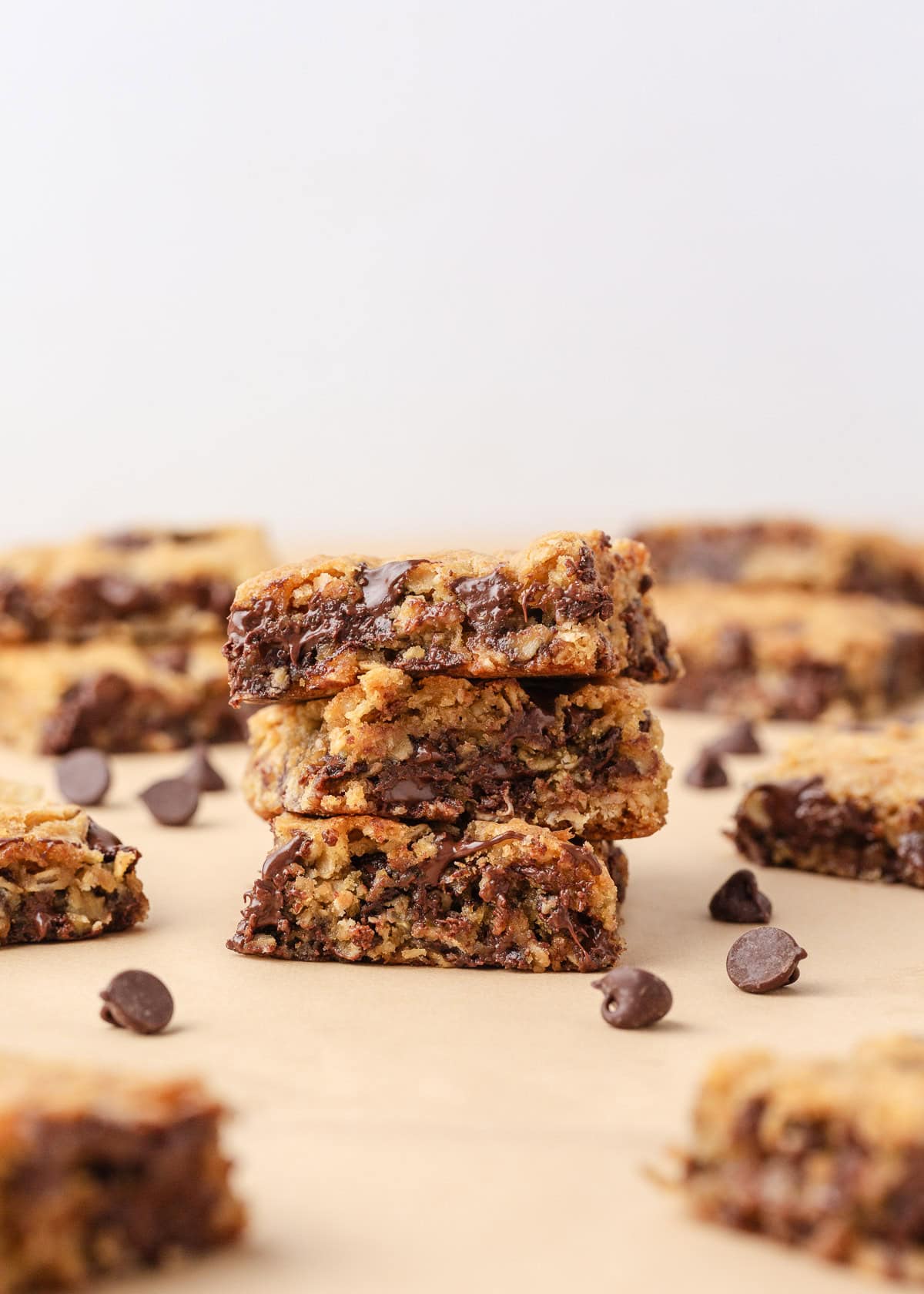 Loaded with oats, brown sugar, and melty chocolate chips, these oatmeal cookie bars are thick, chewy, and made in a single batch. No cookie scoop required! Chewy oatmeal chocolate chip cookie bars stacked on a baking surface with chocolate chips scattered around.