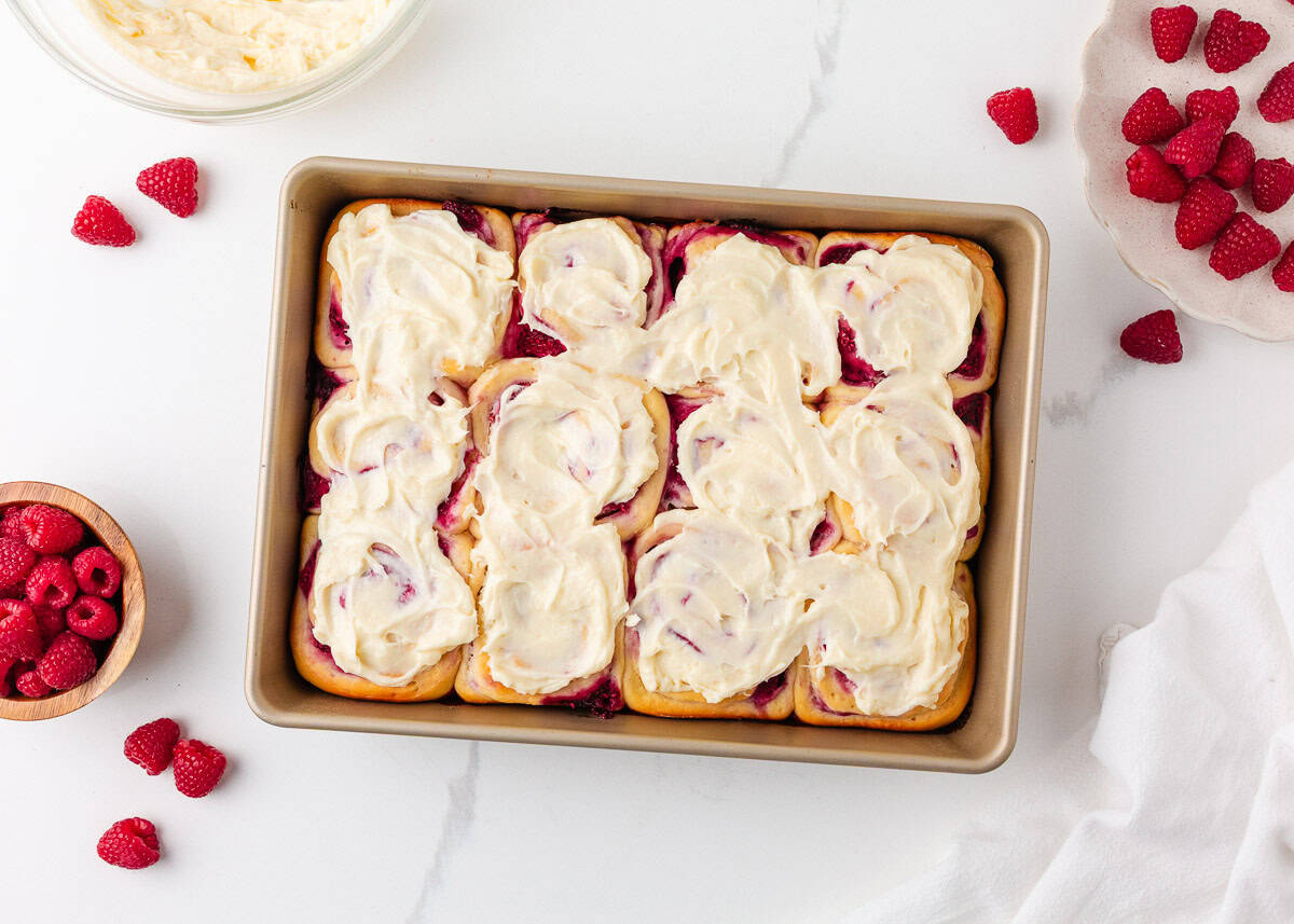 These raspberry sweet rolls are an indulgent breakfast treat made with soft yeast dough, a swirled frozen raspberry filling, and thick cream cheese frosting! Frosted raspberry swirl sweet rolls in baking pan with fresh raspberry and cream cheese icing on counter.