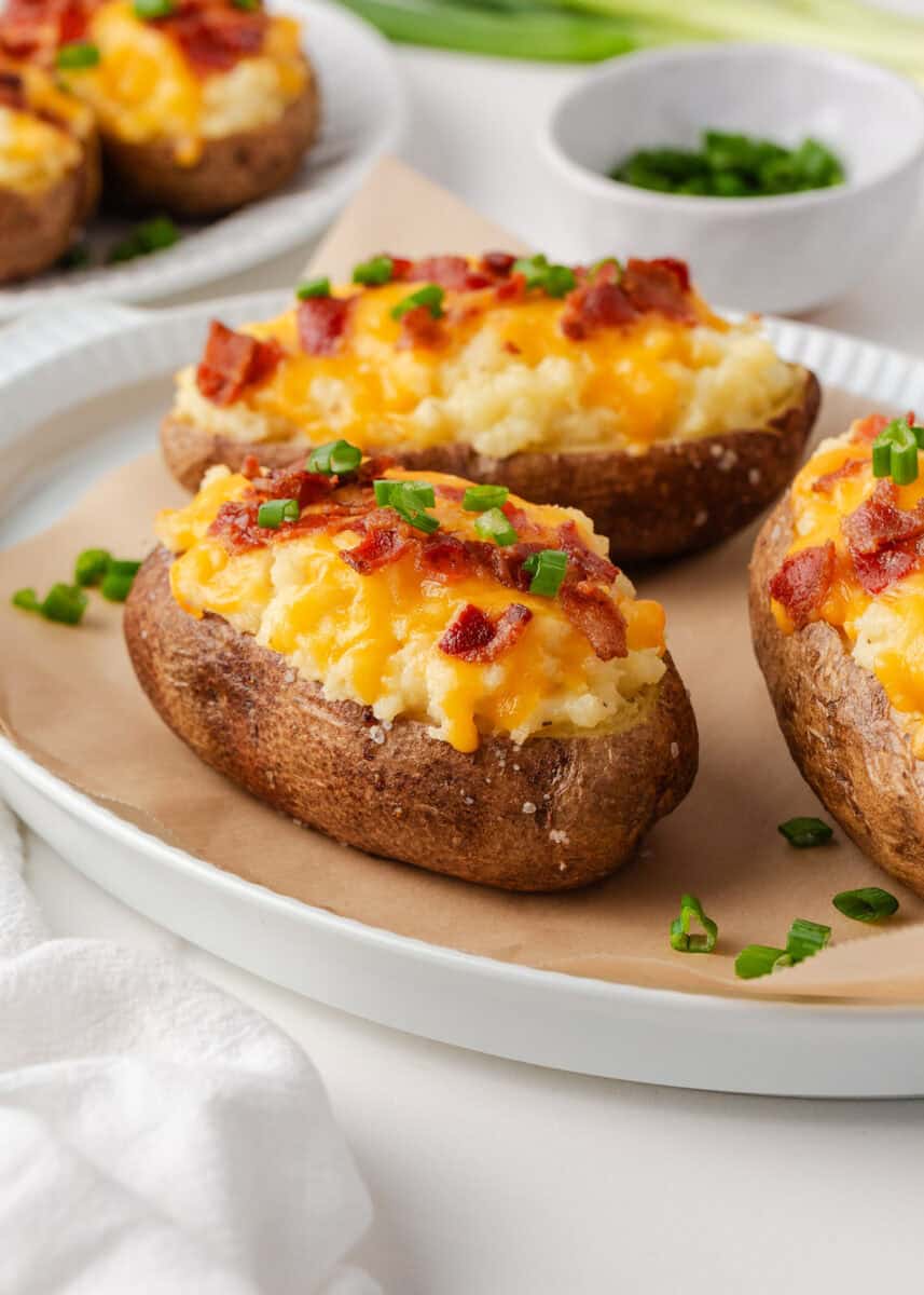The BEST Twice Baked Potatoes Recipe - One of my all-time favorite side dishes! These twice baked potatoes make the perfect appetizer and are a true crowd pleaser! So easy and yummy! Twice baked potatoes topped with melted cheddar cheese, crispy bacon bits, and fresh green onions on a white serving plate.