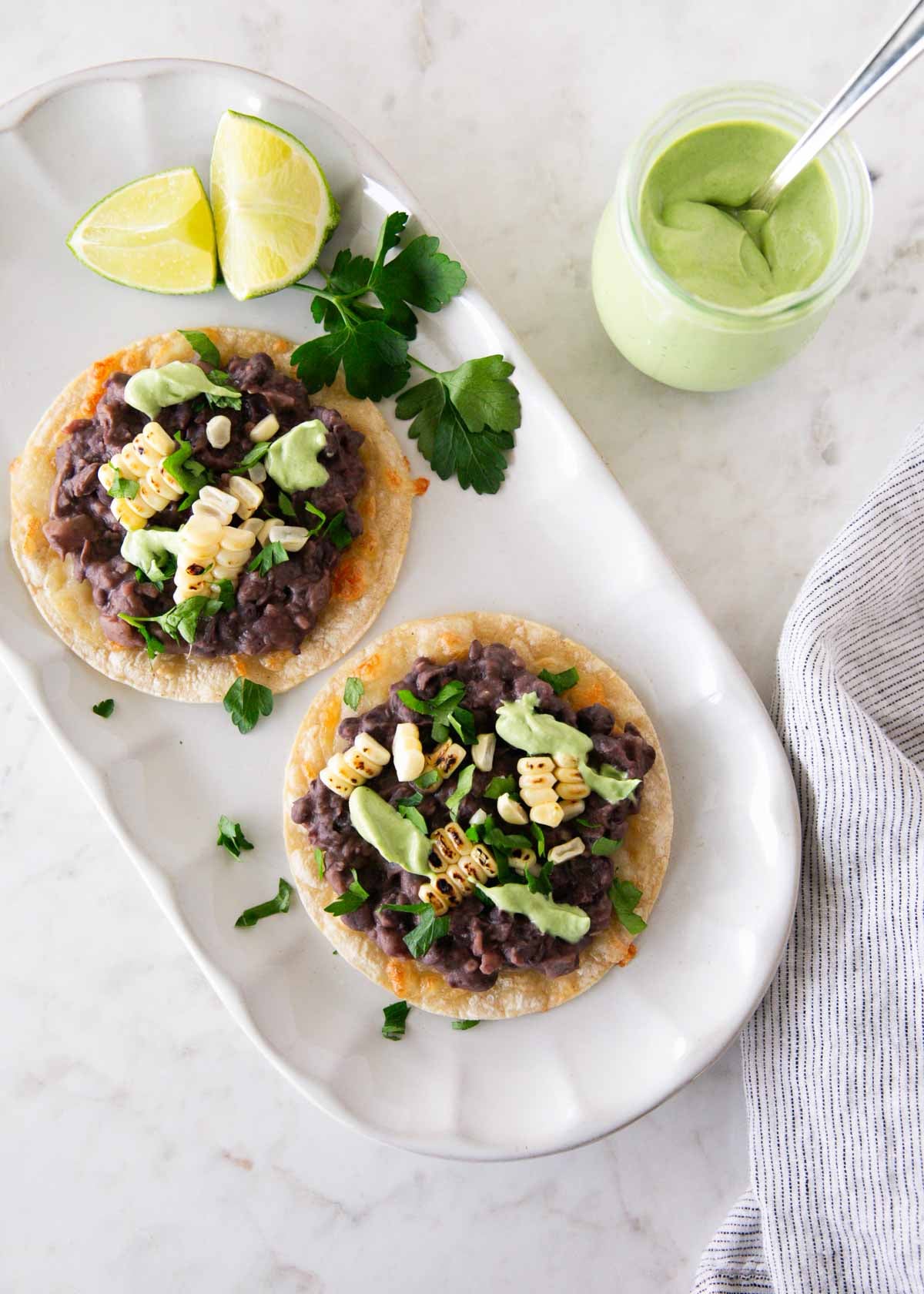 These black bean tostadas start with crispy shells topped with seasoned beans, salsa, grilled corn, and cheese, then finished with homemade avocado crema. Black bean tostadas topped with charred corn, cilantro, and avocado cream on a white platter with lime wedges.