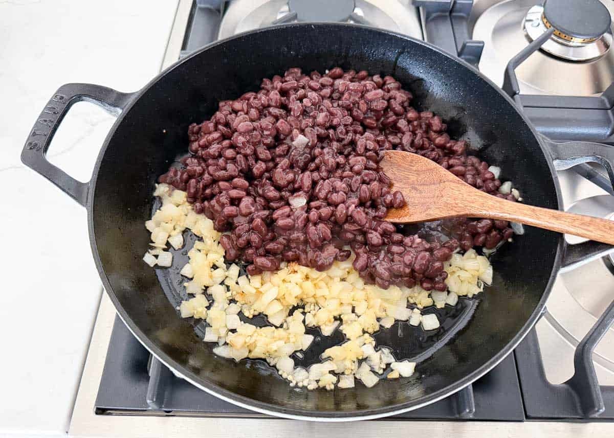 These black bean tostadas start with crispy shells topped with seasoned beans, salsa, grilled corn, and cheese, then finished with homemade avocado crema. Black bean and onions in a skillet.
