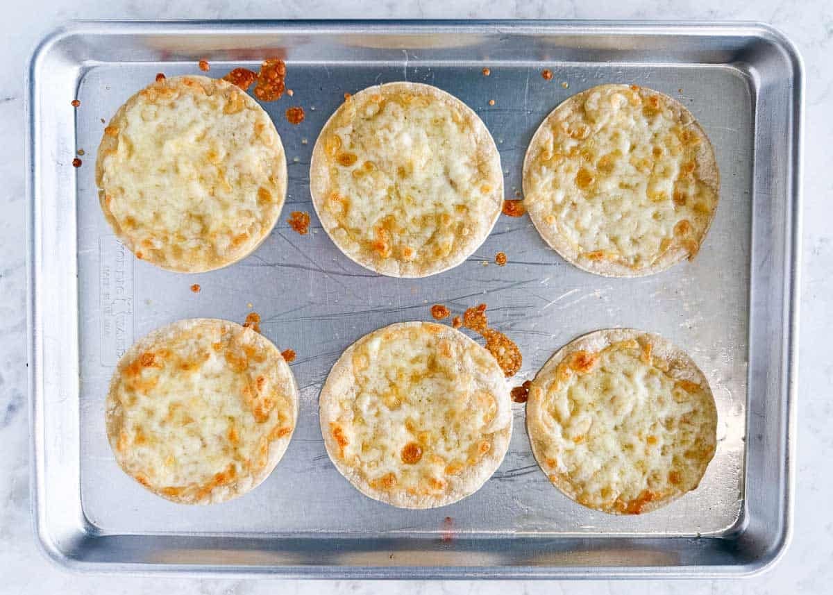 These black bean tostadas start with crispy shells topped with seasoned beans, salsa, grilled corn, and cheese, then finished with homemade avocado crema. Corn tortillas with melted cheese baked on a sheet pan for tostadas.