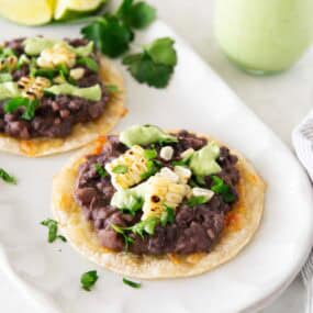 Two black bean tostadas with corn and avocado crema on a serving platter, with a small jar of avocado cream and lime wedges.