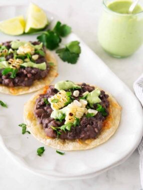 Two black bean tostadas with corn and avocado crema on a serving platter, with a small jar of avocado cream and lime wedges.