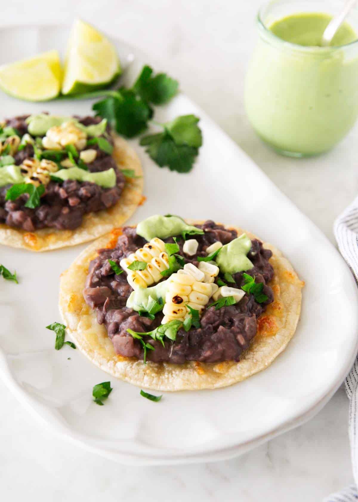 These black bean tostadas start with crispy shells topped with seasoned beans, salsa, grilled corn, and cheese, then finished with homemade avocado crema. Close up of black bean tostadas topped with roasted corn and cilantro, drizzled with creamy avocado sauce.