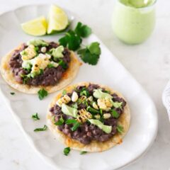 Two black bean tostadas with corn and avocado crema on a serving platter, with a small jar of avocado cream and lime wedges.