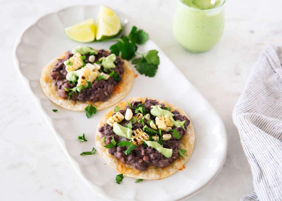 Two black bean tostadas with corn and avocado crema on a serving platter, with a small jar of avocado cream and lime wedges.