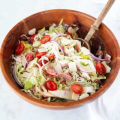 Grinder salad recipe in a wooden bowl with serving spoon, made with lettuce, turkey, salami, provolone, tomatoes, and red onion.