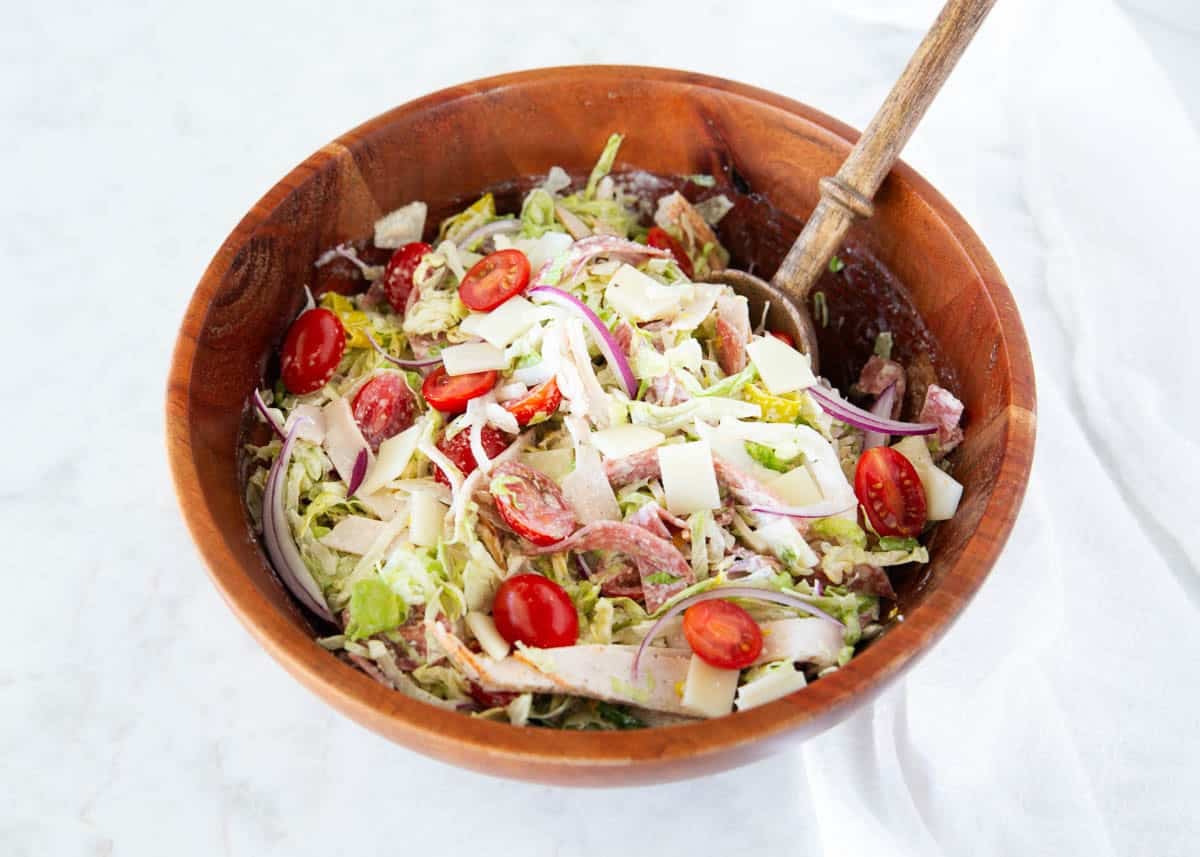 Grinder salad recipe in a wooden bowl with serving spoon, made with lettuce, turkey, salami, provolone, tomatoes, and red onion.