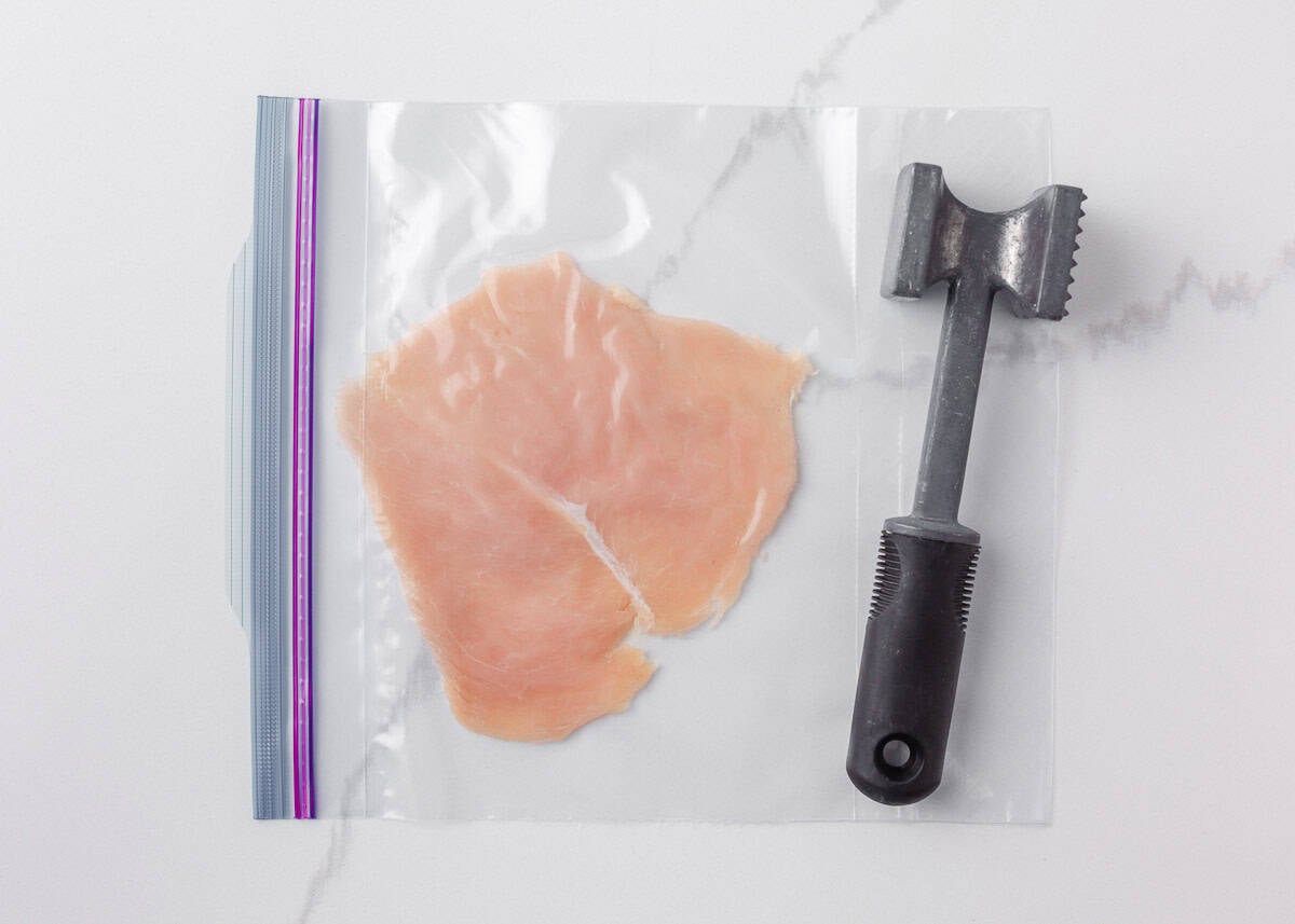 This chicken schnitzel is coated in seasoned breadcrumbs and pan fried until perfectly crispy, then finished with a squeeze of lemon. Thin, juicy, and ready in 30 minutes! Chicken breast in a zip top bag being pounded thin with a meat mallet for chicken schnitzel.