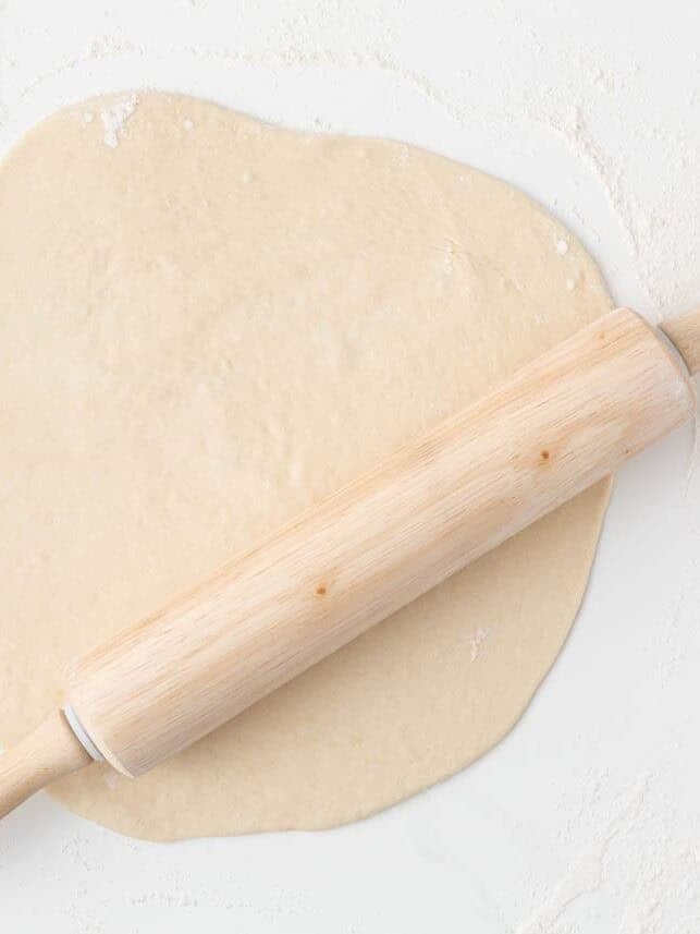 Homemade pizza dough rolled into a round on a floured surface with a wooden rolling pin.