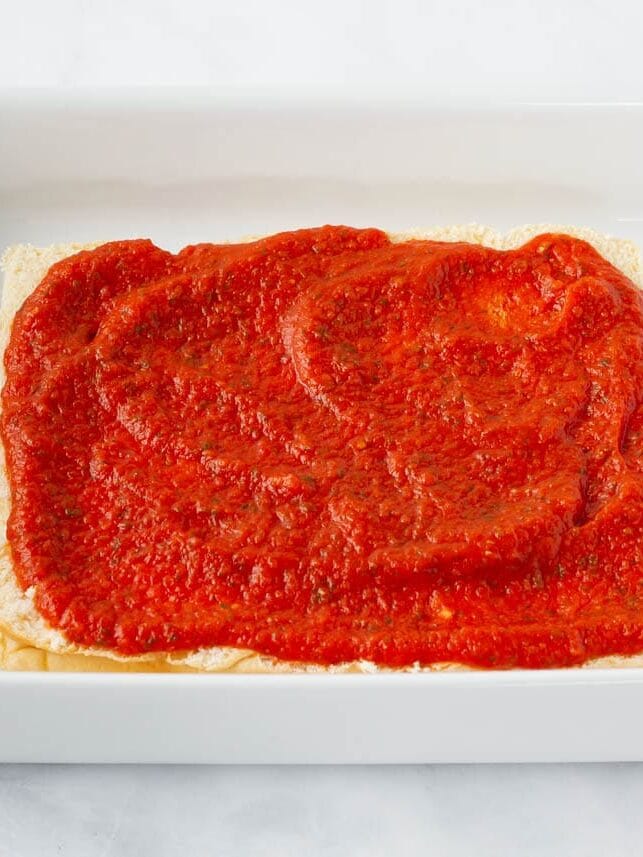 Pizza sauce spread evenly over the bottom half of Hawaiian rolls in a baking dish.
