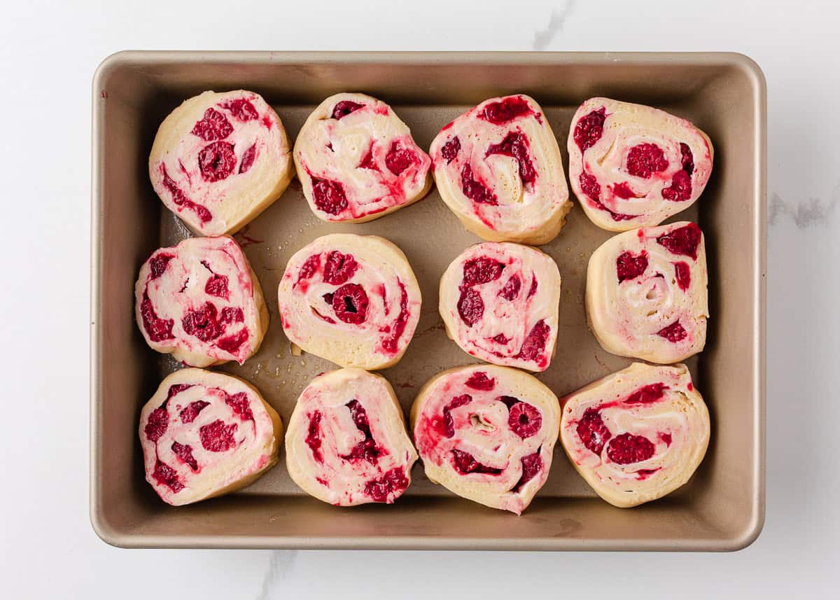 These raspberry sweet rolls are an indulgent breakfast treat made with soft yeast dough, a swirled frozen raspberry filling, and thick cream cheese frosting! Unbaked raspberry sweet rolls sliced and arranged in baking pan before 2nd rise.