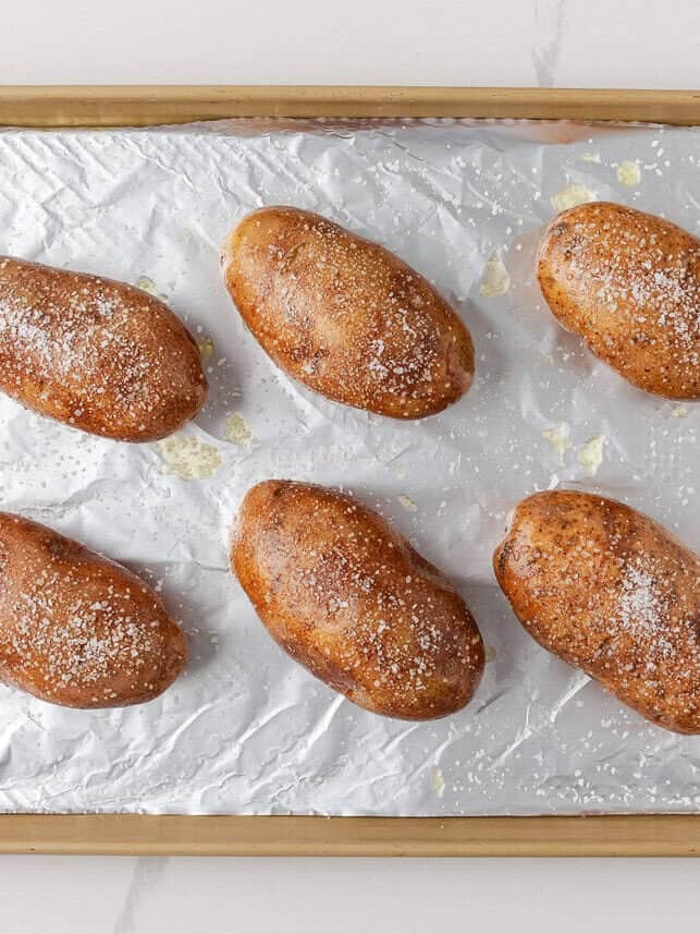 Whole baked russet potatoes brushed with olive oil and sprinkled with salt on a foil lined baking sheet.