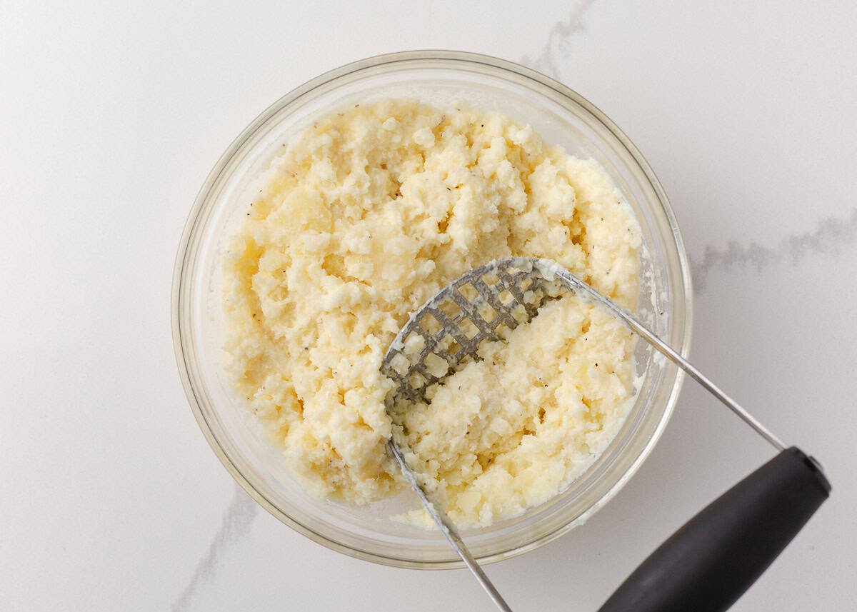 Mashed potato filling being mixed in a glass bowl using a potato masher until smooth and creamy.