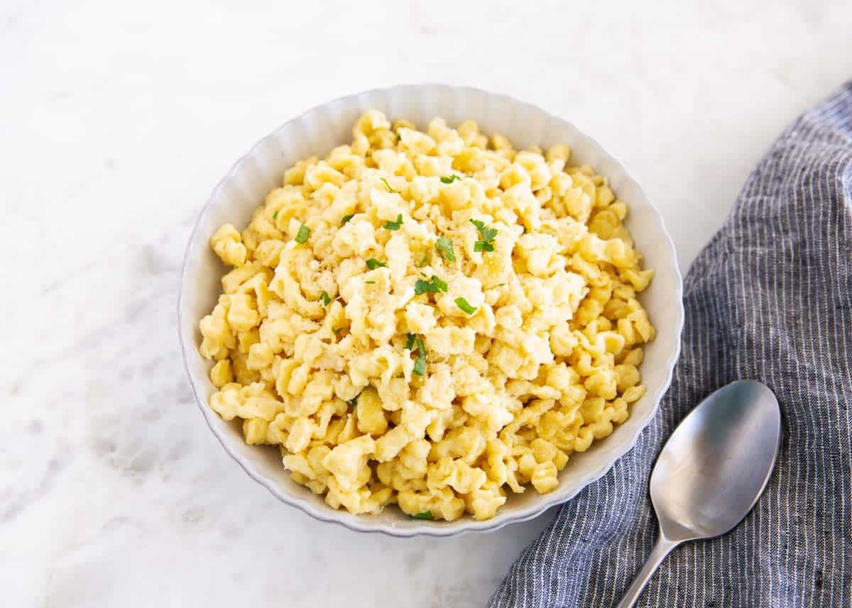 Learn to make homemade German spaetzle that's soft, chewy, and on the table in under 30 minutes. So good with brown butter, gravy, or cheese. Bowl of German spaetzle topped with parmesan and parsley, served beside a spoon and kitchen towel.