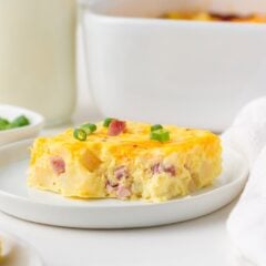 A straight-on view of a square slice of Amish breakfast casserole on a white plate, showing layers of egg, ham, and potato topped with melted cheddar and sliced green onions.
