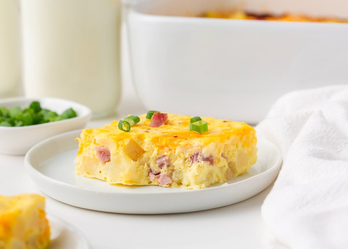 A straight-on view of a square slice of Amish breakfast casserole on a white plate, showing layers of egg, ham, and potato topped with melted cheddar and sliced green onions.