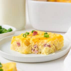 A square slice of Amish breakfast casserole garnished with sliced green onions on a white plate.