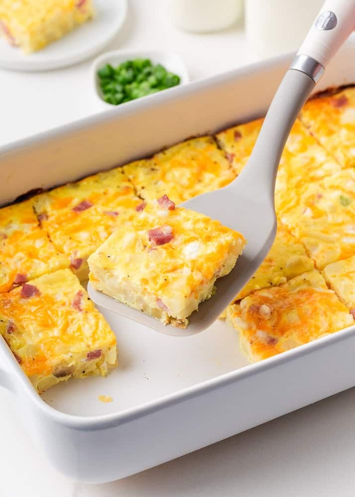 A spatula lifting a square slice of baked breakfast casserole from a white baking dish, showing the egg, ham, potato, and melted cheese layers.