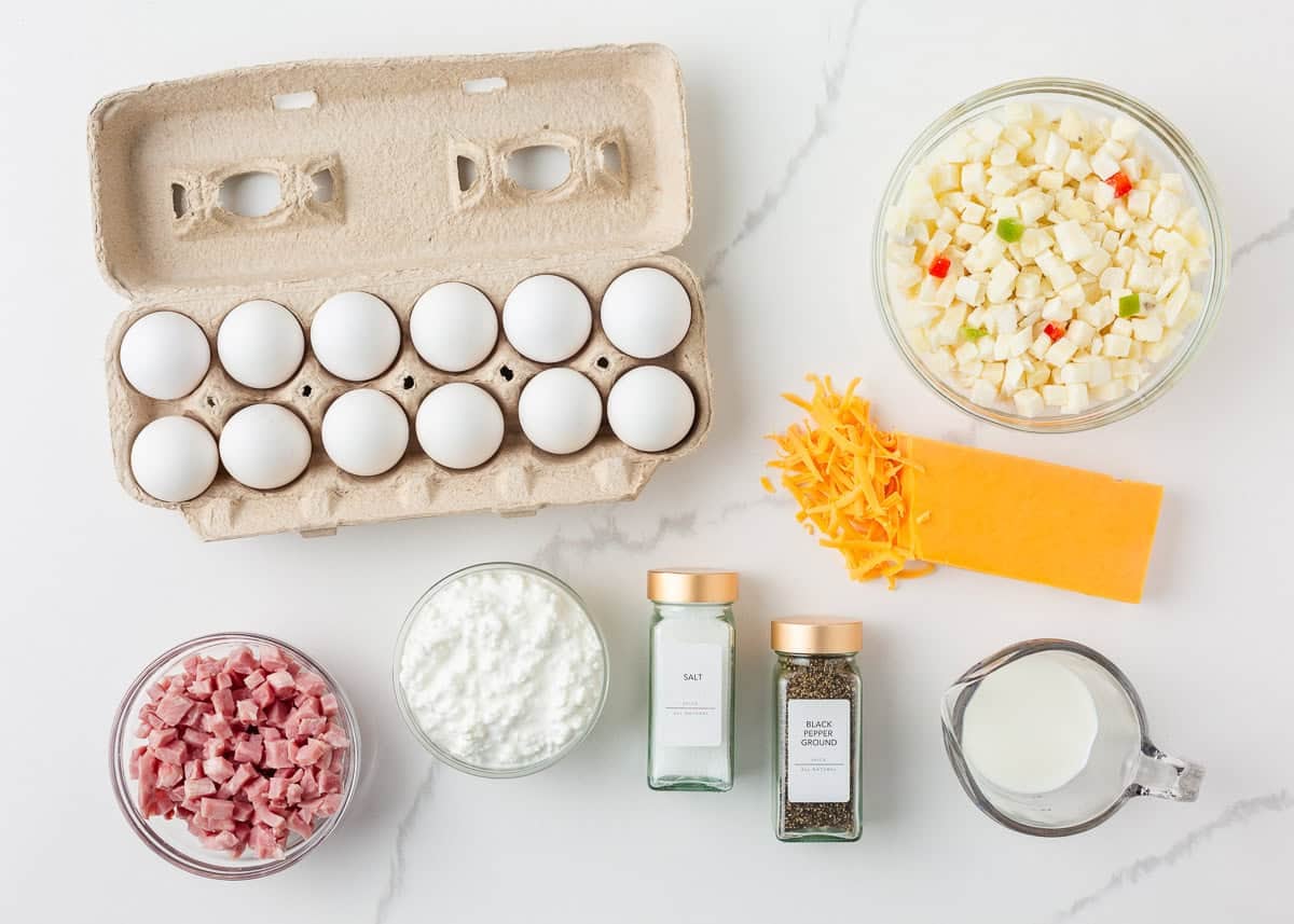 Ingredients for Amish breakfast casserole arranged on a white marble surface, including a carton of 12 eggs, a bowl of frozen potatoes O'Brien, shredded and block cheddar cheese, diced ham, small curd cottage cheese, salt, black pepper, and a glass measuring cup of milk.