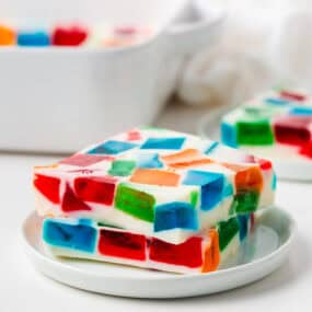 Two thick slices of broken glass jello stacked on a small white plate, showcasing layers of colorful red, blue, green, and orange Jell-O cubes.