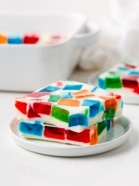 Two thick slices of broken glass jello stacked on a small white plate, showcasing layers of colorful red, blue, green, and orange Jell-O cubes.
