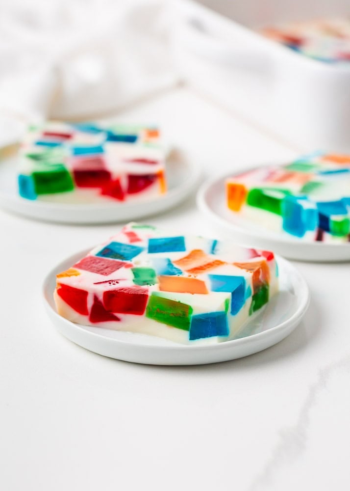 Made with Jello and sweetened condensed milk, stained glass jello has a fun, brightly colored, broken glass look and is the perfect make-ahead dessert for all your parties! A thick square slice of broken glass jello on a small white plate showing layers of colorful Jell-O cubes in red, blue, green, and orange set in a milky white gelatin.