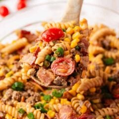 Close-up of a wooden spoon holding a serving of cowboy pasta salad with cherry tomatoes, rotini, ground beef, bacon, and corn in a smoky, creamy dressing.