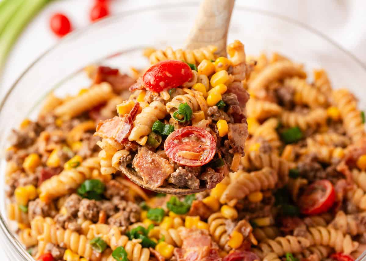 Packed with bold flavors in every bite, this cowboy pasta salad is made with tender noodles, bacon, seasoned ground beef, a creamy dressing, and Southwestern flavor! Close up of a wooden spoon holding a serving of cowboy pasta salad with cherry tomatoes, rotini, ground beef, bacon, and corn in a smoky, creamy dressing.