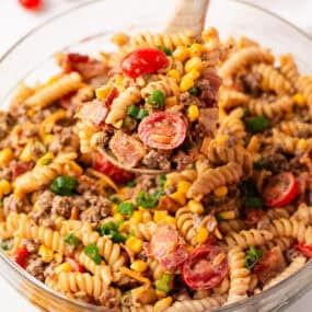 A wooden spoon lifts a heaping scoop of cowboy pasta salad from a large glass bowl, showing rotini, ground beef, bacon, corn, cherry tomatoes, and green onions in a creamy BBQ dressing.