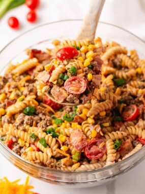 A wooden spoon lifts a heaping scoop of cowboy pasta salad from a large glass bowl, showing rotini, ground beef, bacon, corn, cherry tomatoes, and green onions in a creamy BBQ dressing.