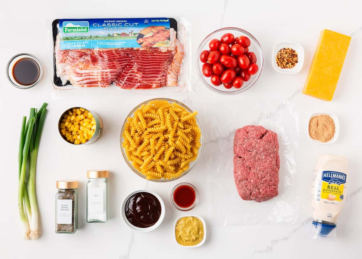 Packed with bold flavors in every bite, this cowboy pasta salad is made with tender noodles, bacon, seasoned ground beef, a creamy dressing, and Southwestern flavor! Flat lay of all the ingredients needed to make cowboy pasta salad, including rotini, ground beef, bacon, cherry tomatoes, corn, cheddar cheese, and condiments.