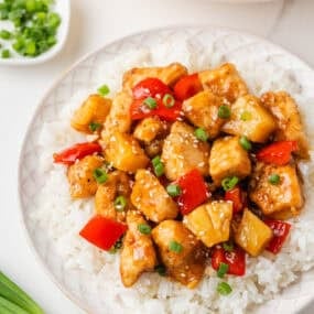 Close-up of pineapple chicken served over white rice on a white plate, garnished with sliced green onions and sesame seeds, with a pan of the dish and extra green onions visible in the background.