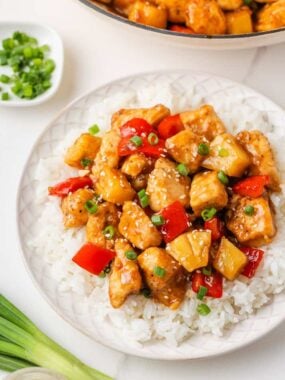 Close-up of pineapple chicken served over white rice on a white plate, garnished with sliced green onions and sesame seeds, with a pan of the dish and extra green onions visible in the background.