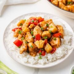 Pineapple chicken stir fry with red bell pepper and pineapple chunks served over fluffy white rice on a white plate, topped with sesame seeds and chopped green onions, with a skillet of the dish in the background.
