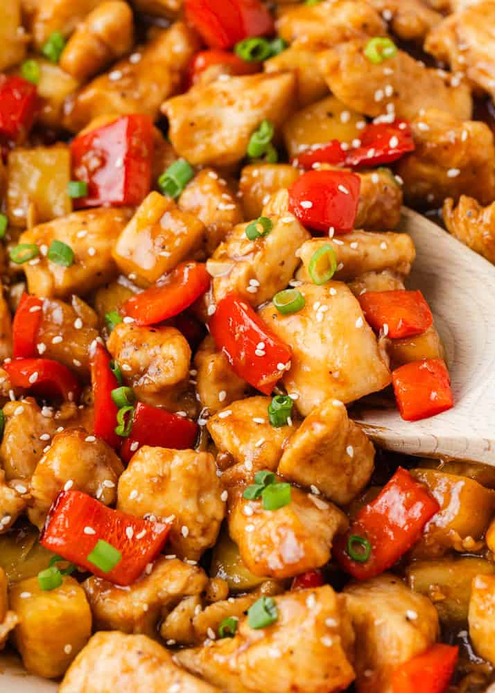 This pineapple chicken is sweet, tangy, and on the table in 30 minutes. Tender chicken, crisp bell peppers, and juicy pineapple in a sauce that beats takeout every time. Close-up of pineapple chicken in a skillet, showing saucy chunks of chicken, red bell pepper, and pineapple tossed in a glossy sauce and garnished with green onions and sesame seeds, with a wooden spoon scooping a serving.