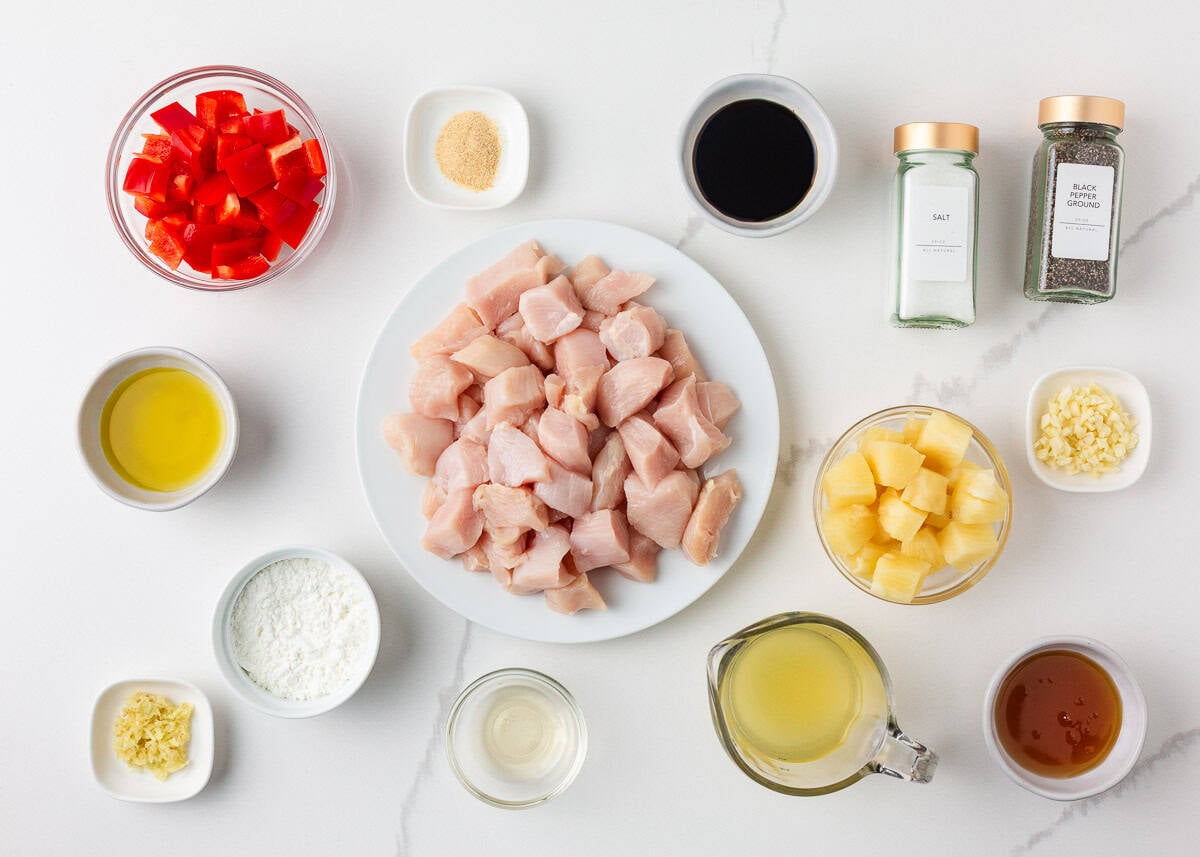 This pineapple chicken is sweet, tangy, and on the table in 30 minutes. Tender chicken, crisp bell peppers, and juicy pineapple in a sauce that beats takeout every time. Overhead view of pineapple chicken ingredients laid out on a white marble surface, including cubed raw chicken breast, chopped red bell pepper, pineapple chunks, soy sauce, pineapple juice, olive oil, cornstarch, garlic, ginger, honey, salt, and black pepper.