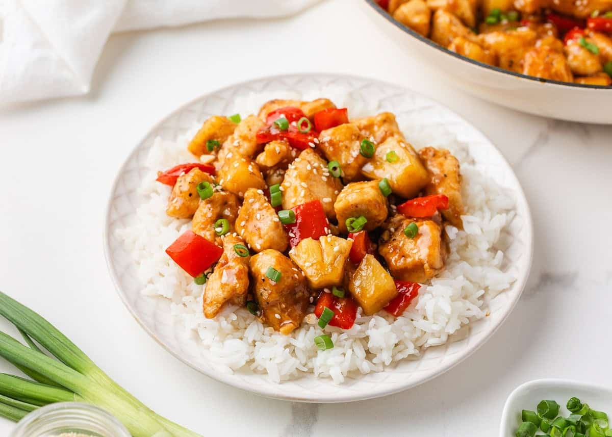 This pineapple chicken is sweet, tangy, and on the table in 30 minutes. Tender chicken, crisp bell peppers, and juicy pineapple in a sauce that beats takeout every time. Pineapple chicken stir fry with red bell pepper and pineapple chunks served over fluffy white rice on a white plate, topped with sesame seeds and chopped green onions, with a skillet of the dish in the background.