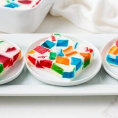 Three servings of colorful stained glass jello arranged on a white rectangular platter, displaying the mosaic-like pattern of jewel-toned Jell-O pieces held together by creamy white gelatin.