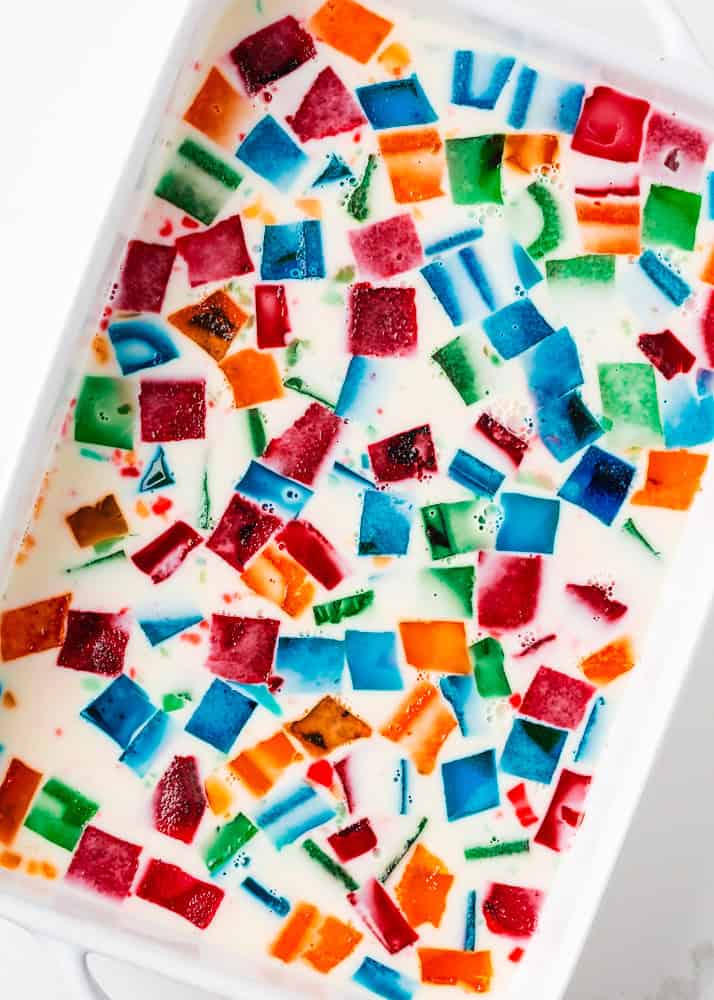 Made with Jello and sweetened condensed milk, stained glass jello has a fun, brightly colored, broken glass look and is the perfect make-ahead dessert for all your parties! Overhead view of a white ceramic baking dish filled with stained glass jello featuring scattered cubes of multicolored Jell-O floating in a sweet condensed milk gelatin mixture.