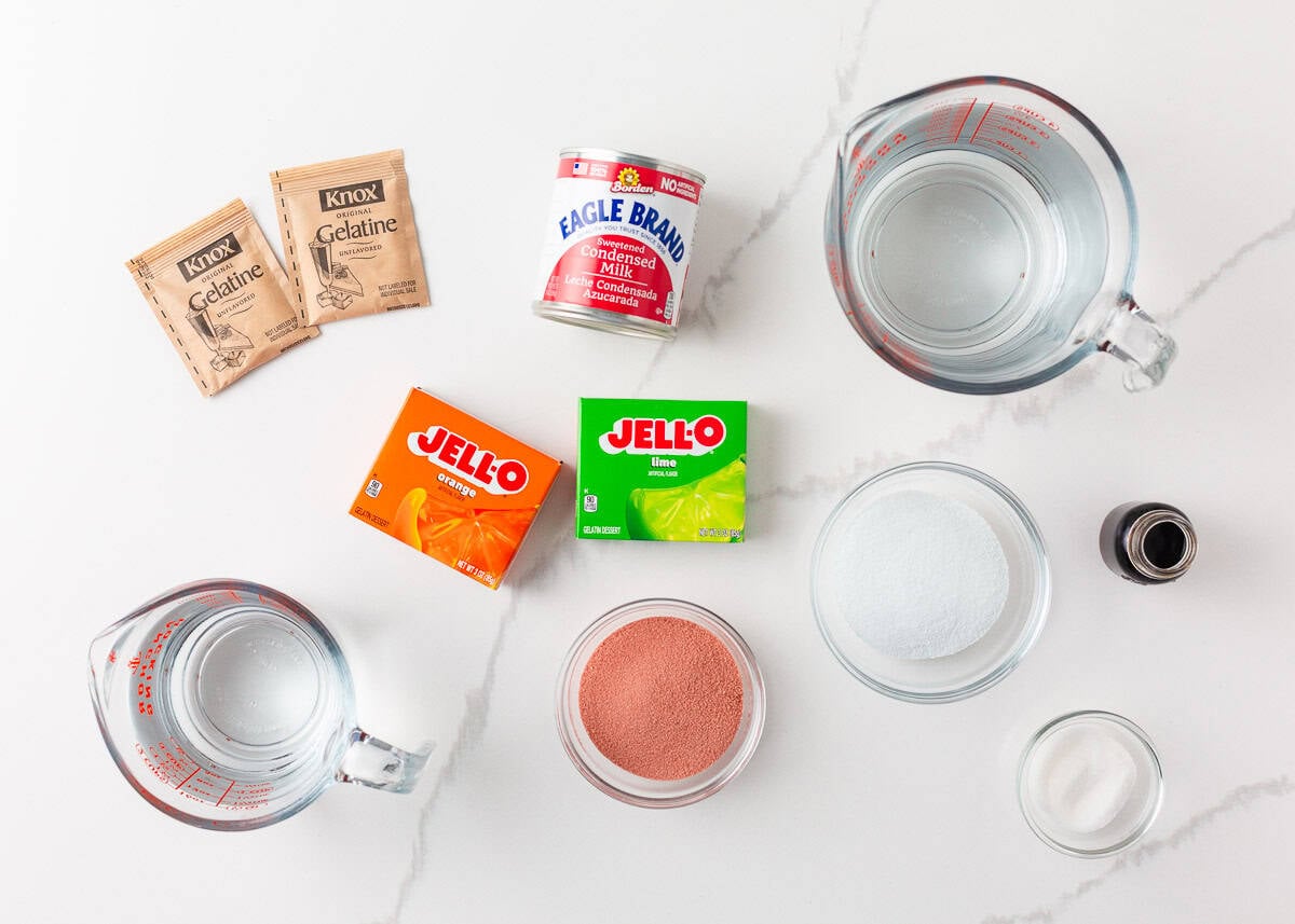 Made with Jello and sweetened condensed milk, stained glass jello has a fun, brightly colored, broken glass look and is the perfect make-ahead dessert for all your parties! Stained glass jello ingredients laid out on a marble countertop including Knox gelatin packets, sweetened condensed milk, Jell-O orange and lime boxes, a bowl of strawberry Jell-O powder, sugar, vanilla extract, and water in glass measuring cups.