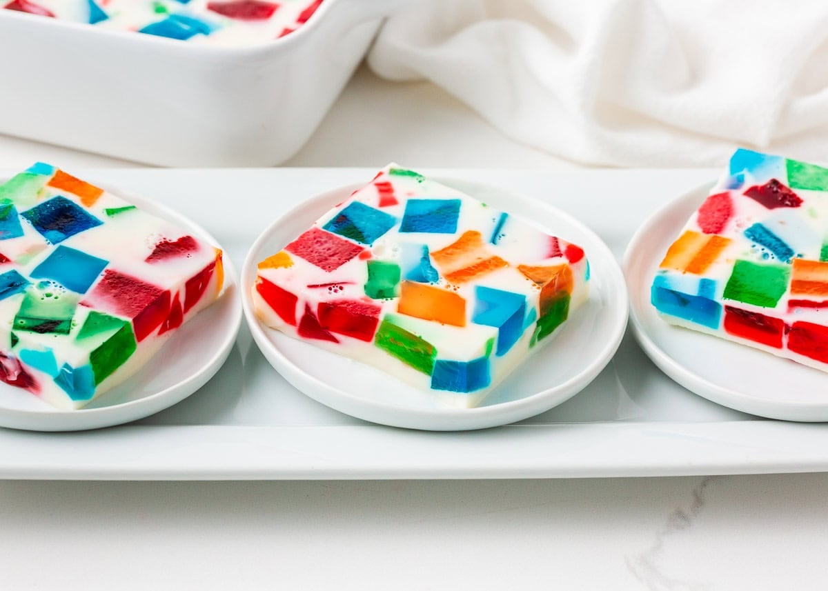 Three servings of rainbow stained glass jello arranged on a white rectangular platter, displaying the mosaic-like pattern of jewel-toned Jell-O pieces held together by creamy white gelatin.