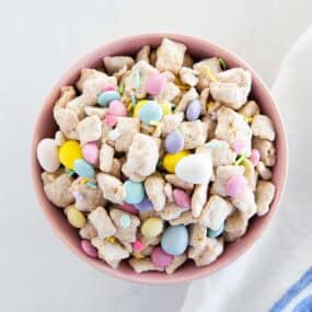 Easter puppy chow snack mix in a pink bowl on a white surface with pastel candies and sprinkles.