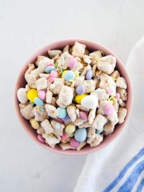 Easter puppy chow snack mix in a pink bowl on a white surface with pastel candies and sprinkles.