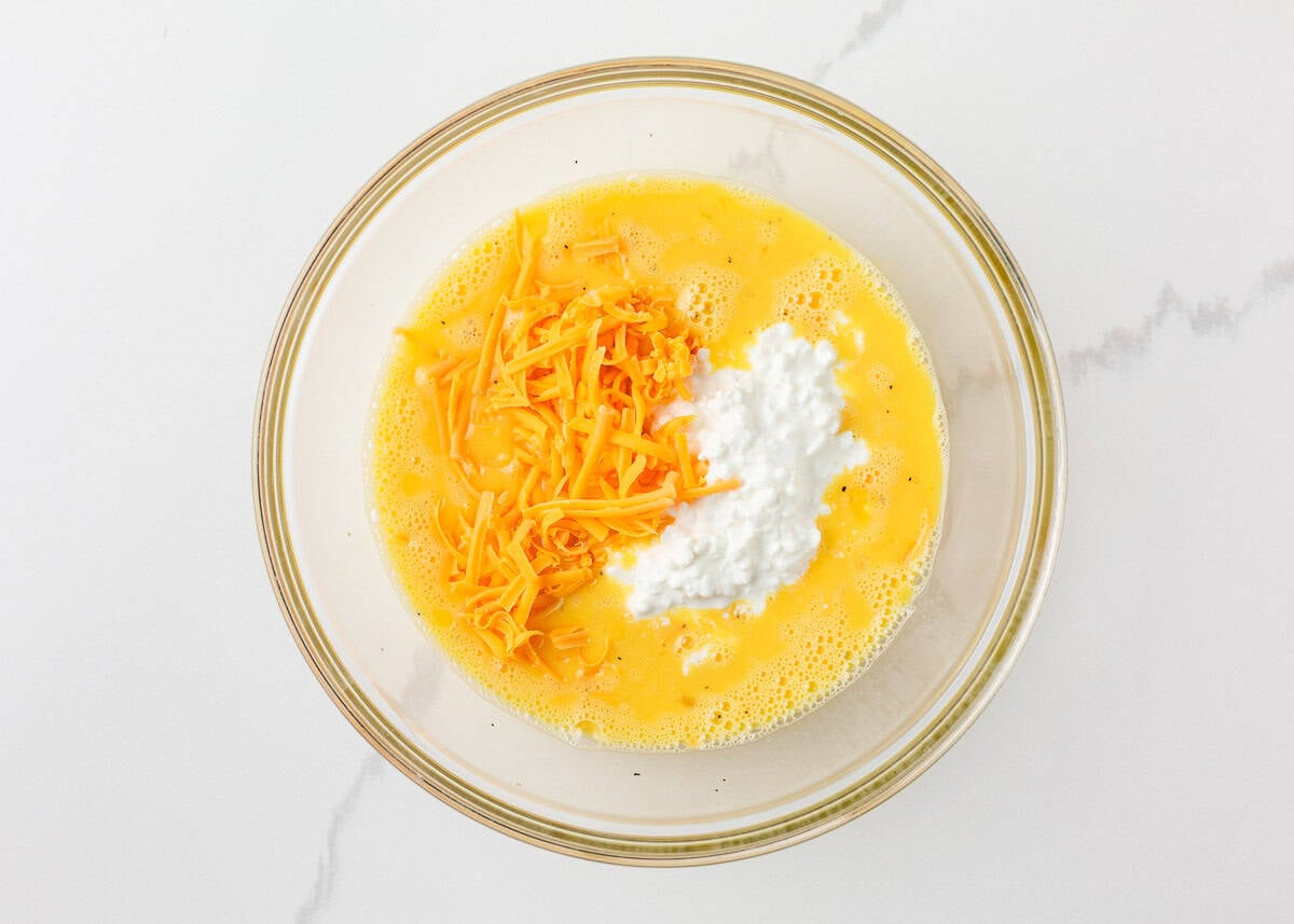 Overhead view of a large glass mixing bowl with whisked eggs and milk, topped with shredded cheddar cheese and a scoop of small curd cottage cheese, before stirring to combine.