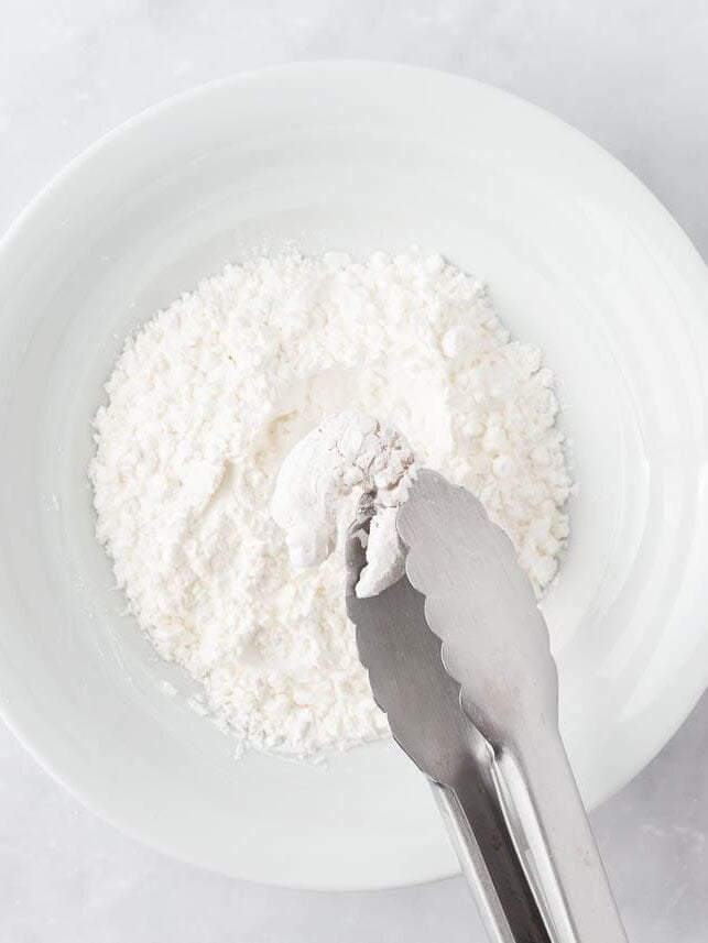 Tongs coating a shrimp in cornstarch in a shallow white bowl.