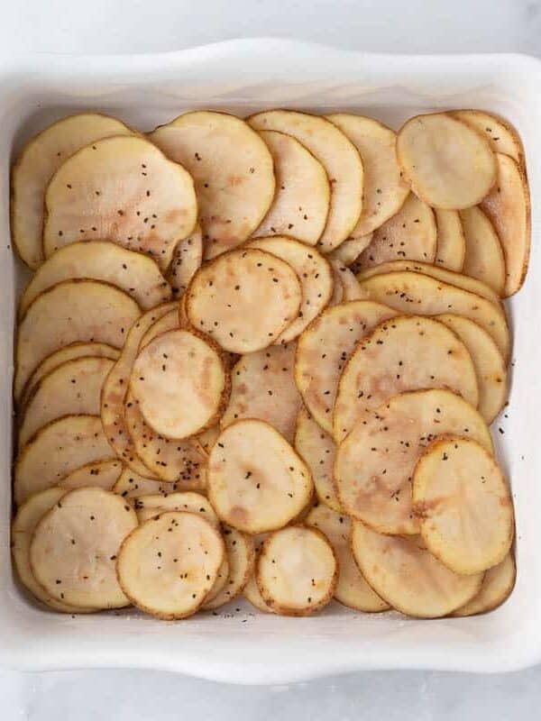 Thinly sliced potatoes layered in a white baking dish seasoned with salt and pepper